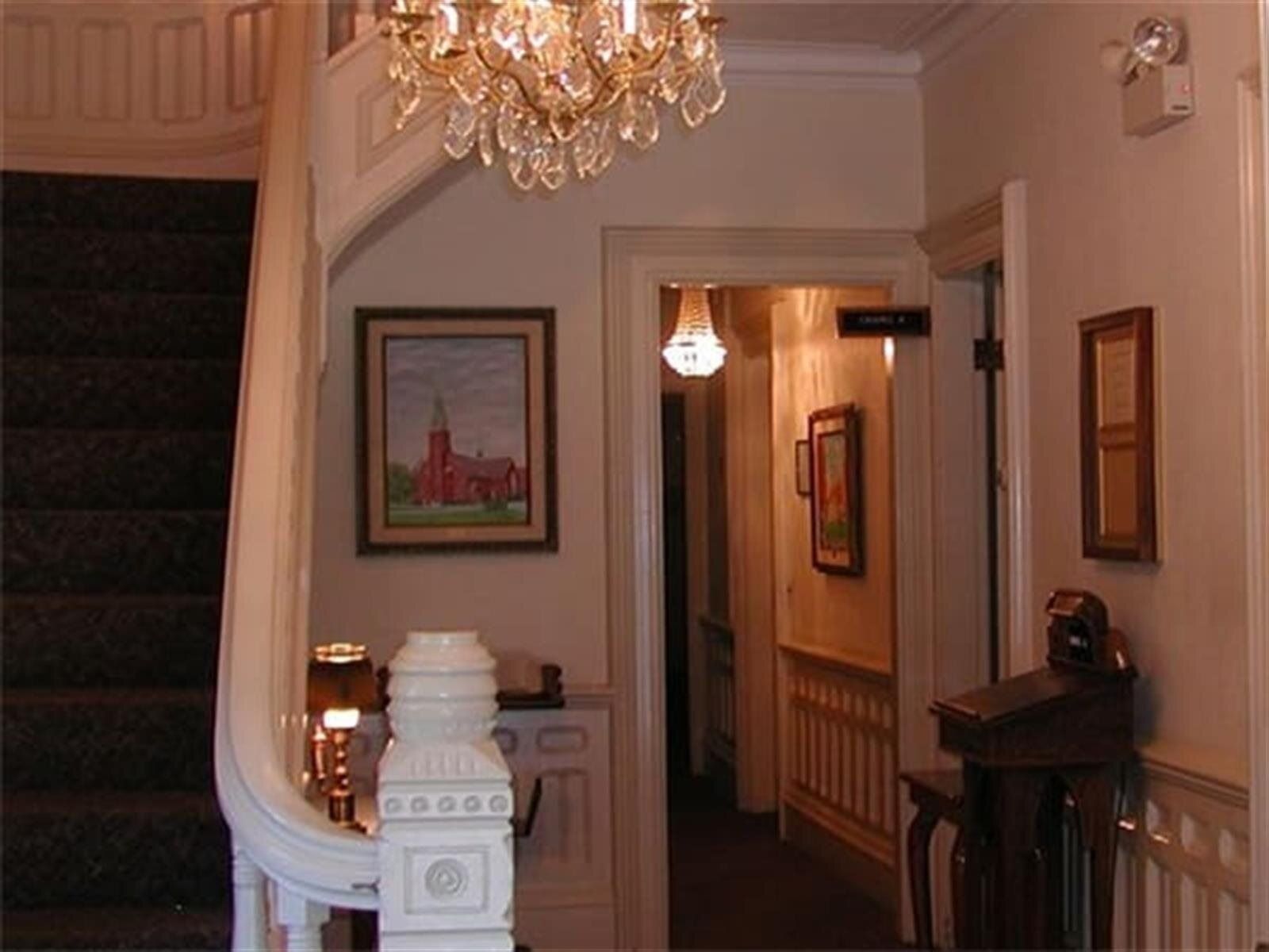 A grand staircase in a home, with a chandelier, art, and hallway visible. Warm lighting and traditional decor.