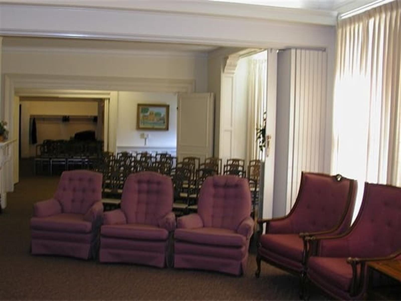 A waiting room with pink upholstered chairs and rows of chairs set up for a service. Light pours in from a window with cream-colored curtains.