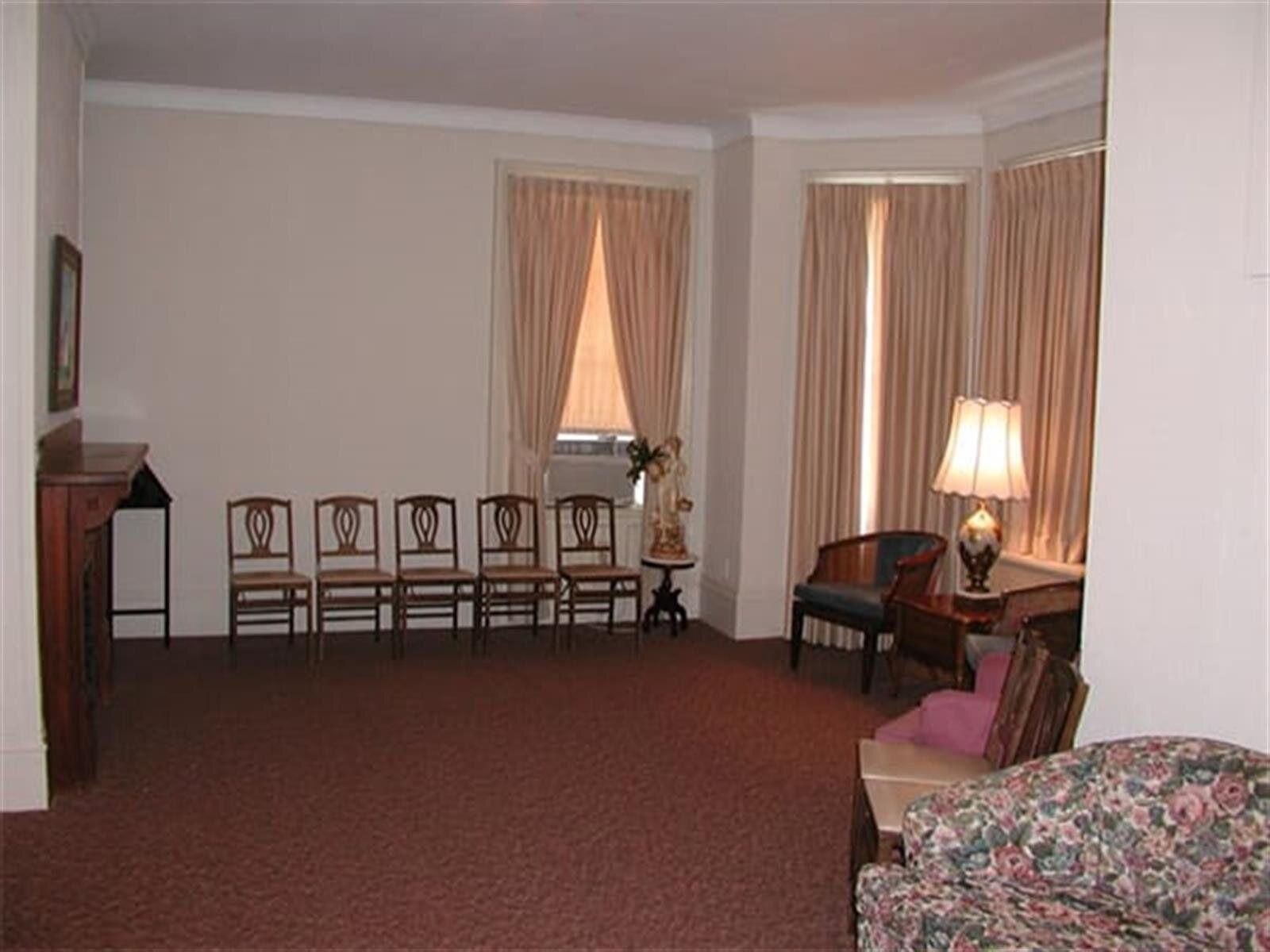 A room with chairs arranged for seating, brown carpet, beige walls, and light-colored curtains; likely a waiting area.