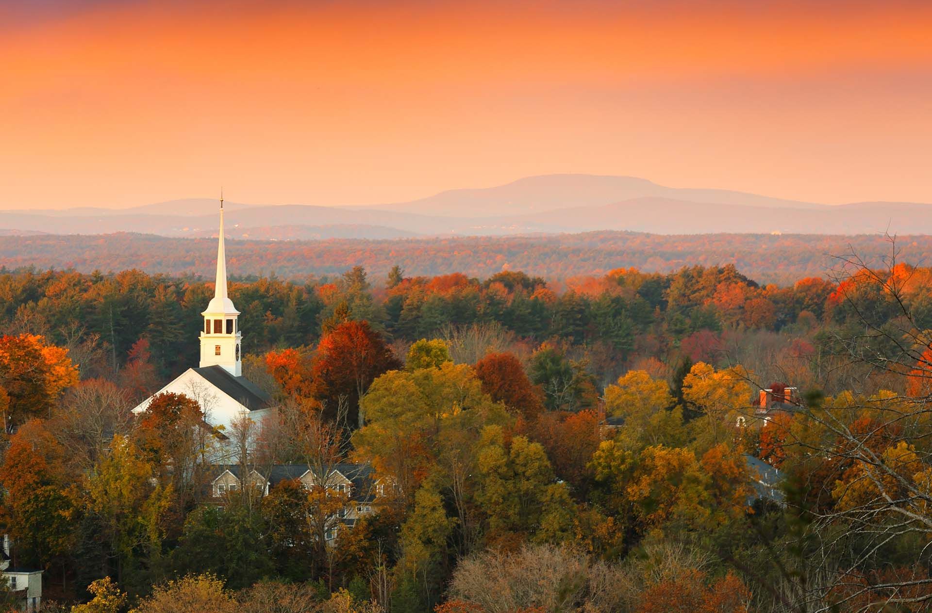A white church with a tall steeple sits amid colorful autumn trees under an orange sky.