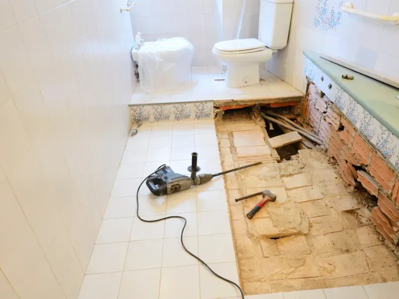A bathroom under renovation with a jackhammer on tiled flooring next to an exposed, unfinished subfloor and brick wall.