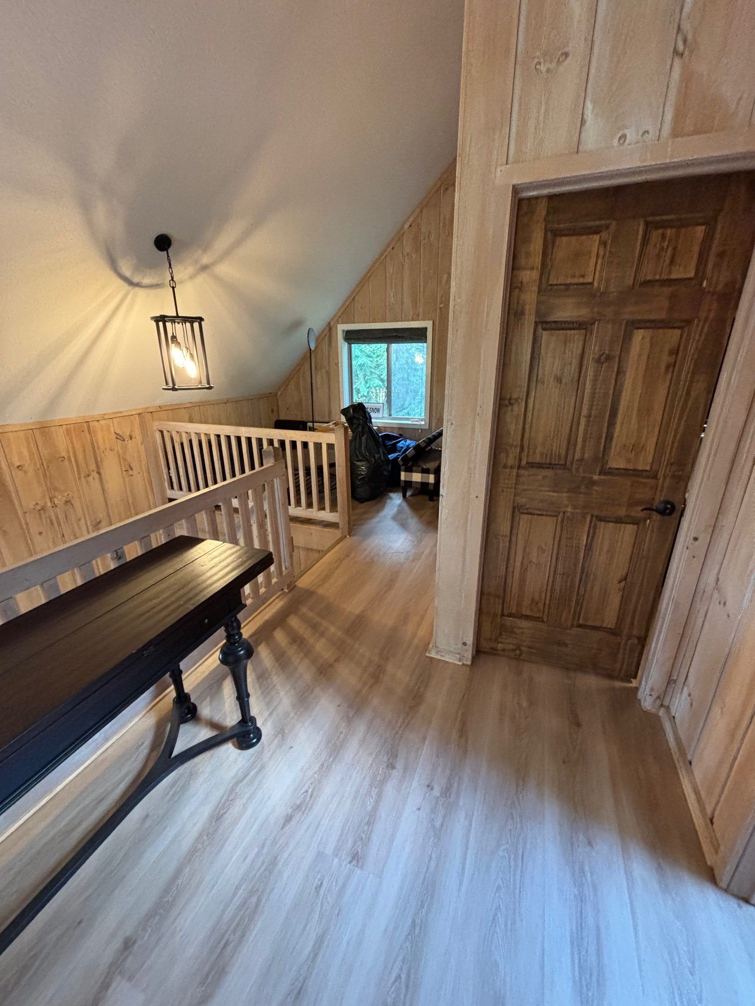 A loft hallway with light wood paneling, a dark wood door, light wood-look flooring, a black console table, and a railing.