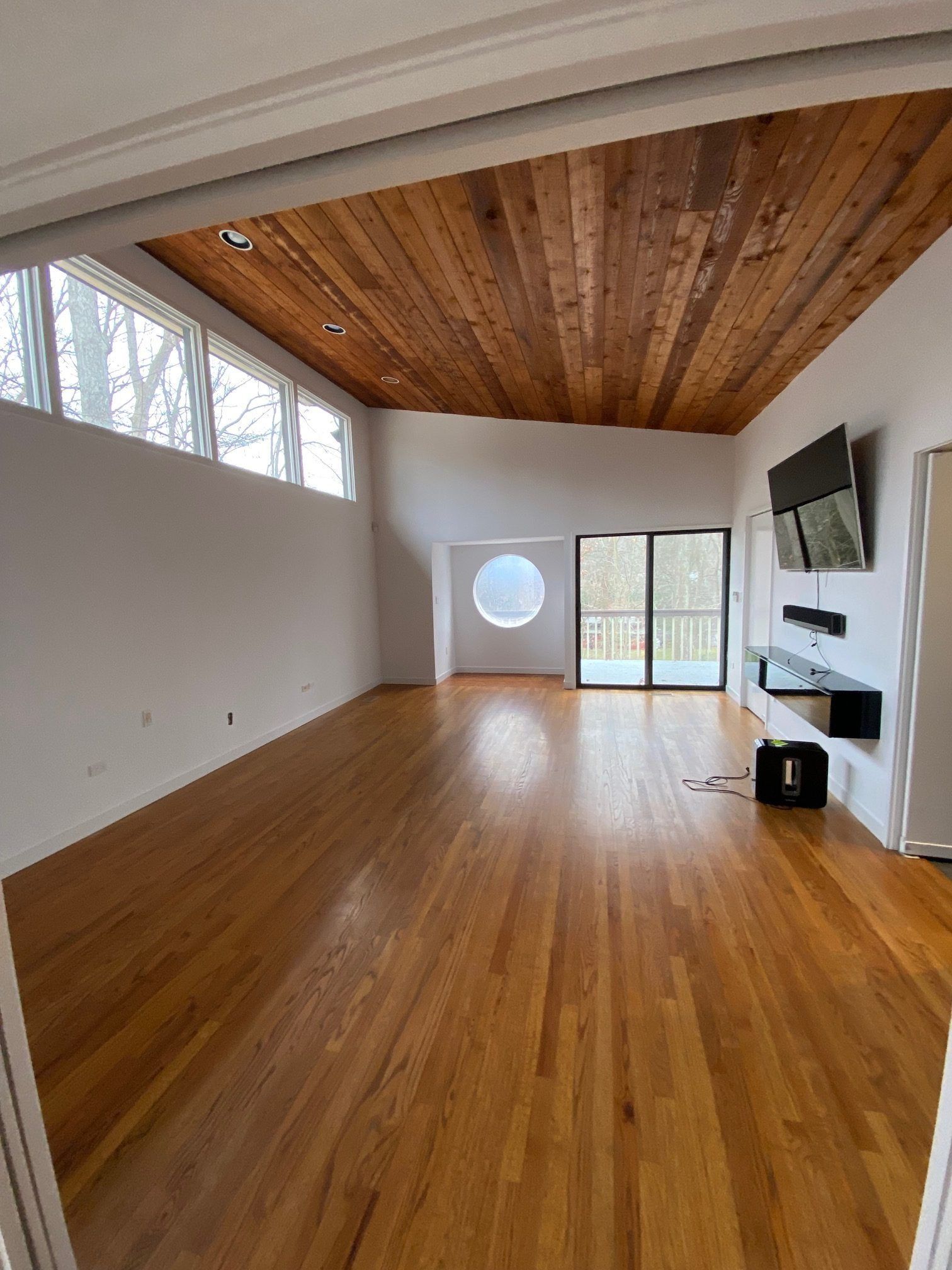 A spacious room with wood floors and a wood-slatted ceiling, featuring a round window, sliding doors, and a wall-mounted TV.
