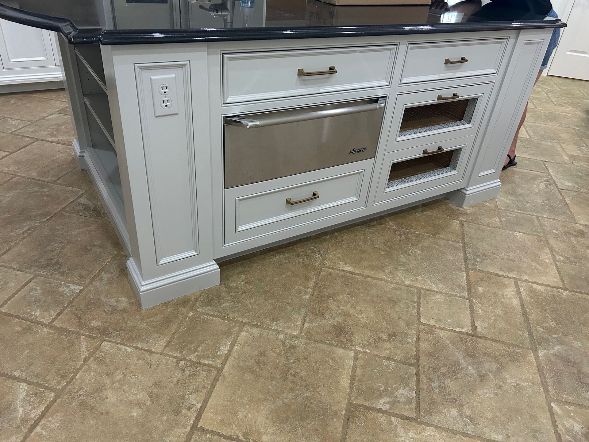 White kitchen island with a stainless steel microwave drawer, drawers, and shelving set on beige stone tile flooring.