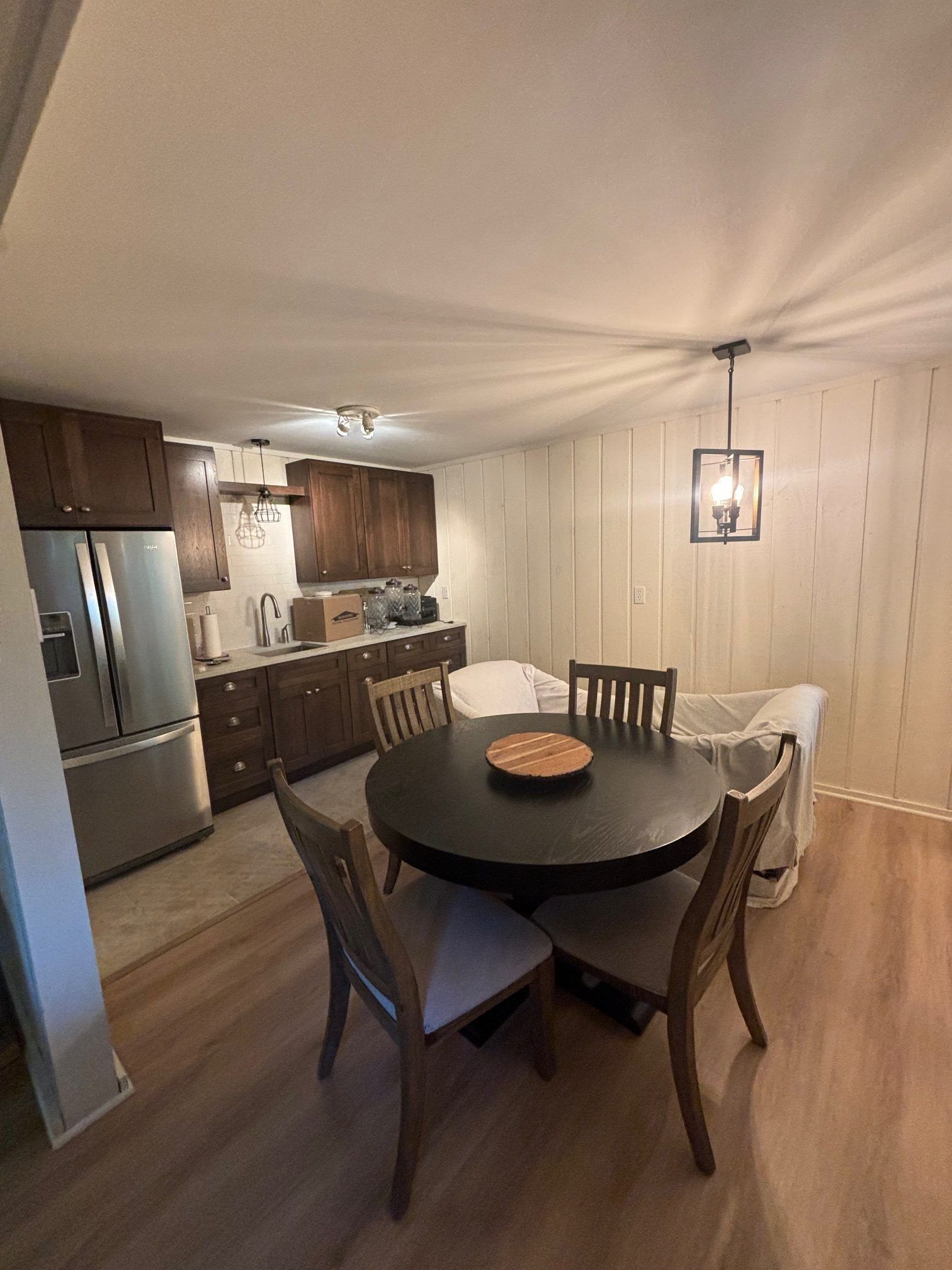 A dining area with a round dark table and chairs, adjacent to a kitchen with dark cabinetry and stainless steel appliances.