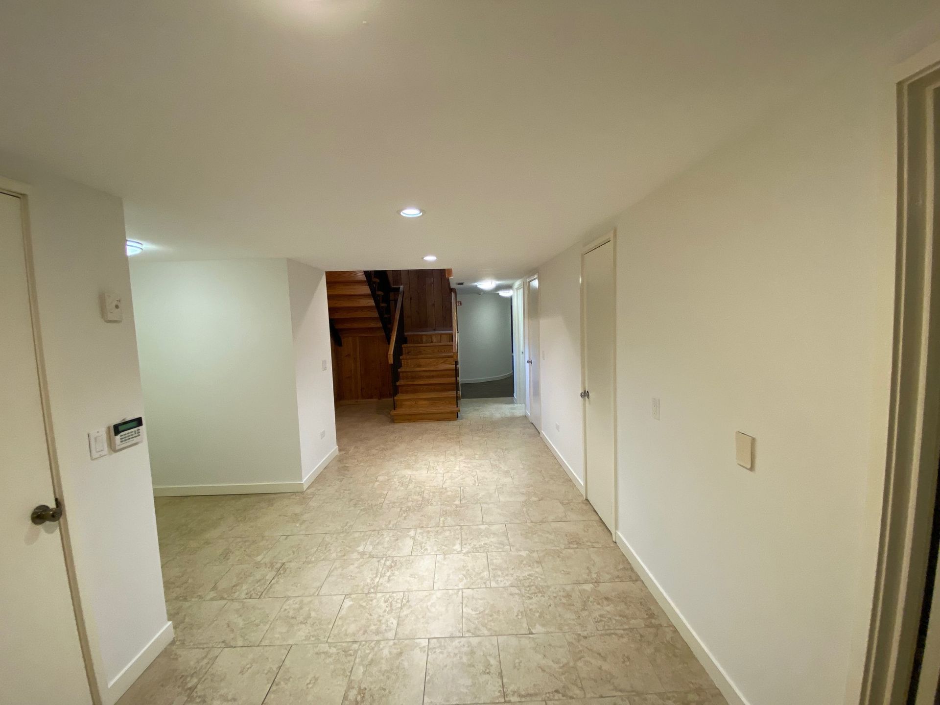 An empty basement hallway with white walls, tiled floor, and wooden stairs leading to a higher level.