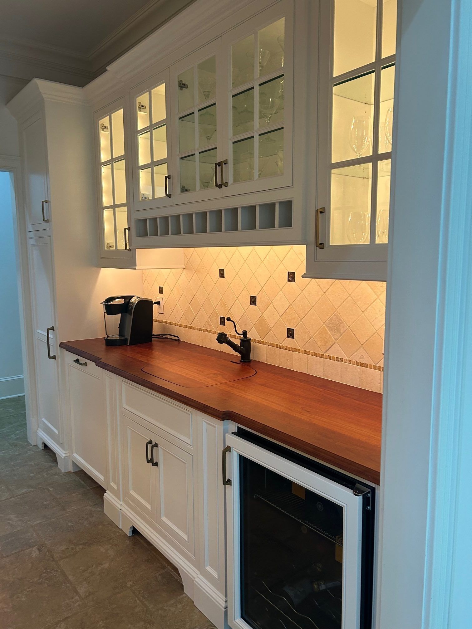 A wet bar with white cabinets, a dark wood countertop, glass upper cabinets, a wine fridge, and a tiled backsplash.
