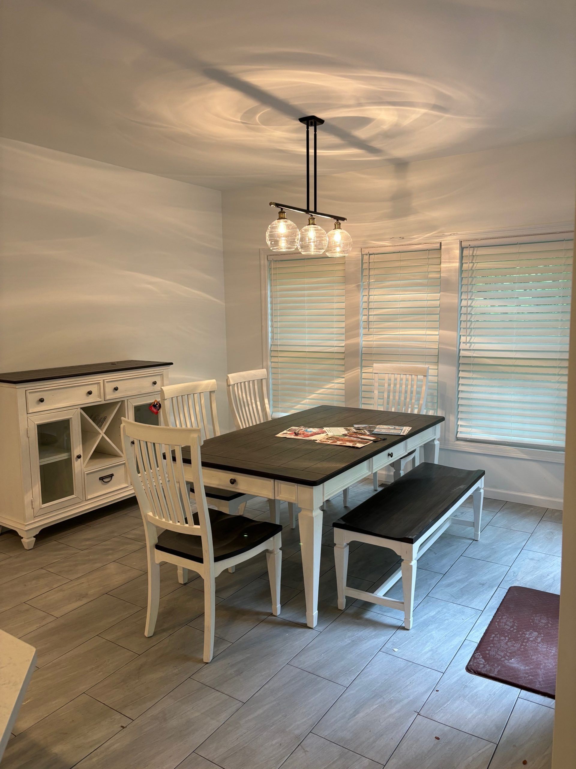 A dining room with white furniture, a dark wood-top table, a matching bench, two chairs, and a three-bulb light fixture.