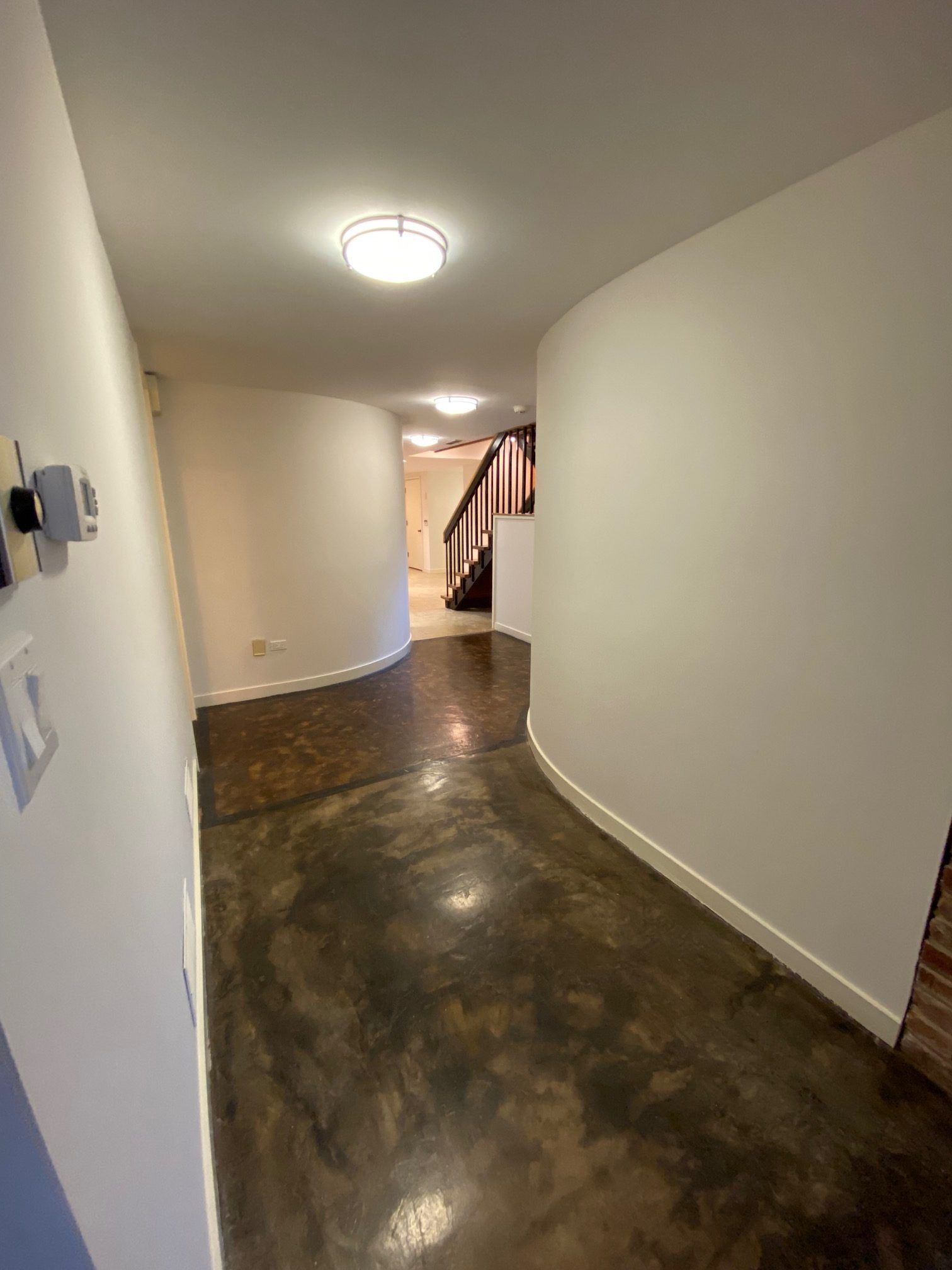 A curved hallway with white walls and a polished, mottled dark brown concrete floor leading toward a staircase.