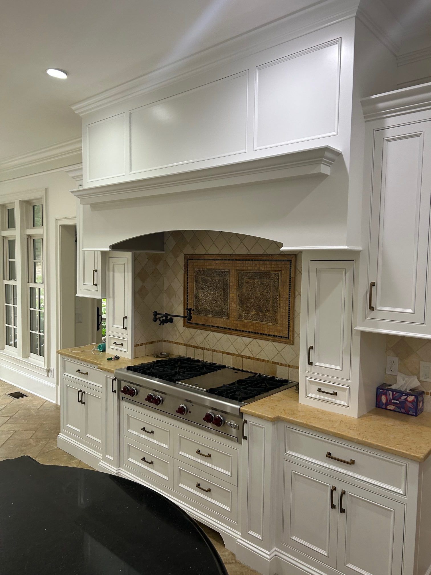 A white custom kitchen hood and range area with cream-colored countertops, beige tile backsplash, and dark metal hardware.