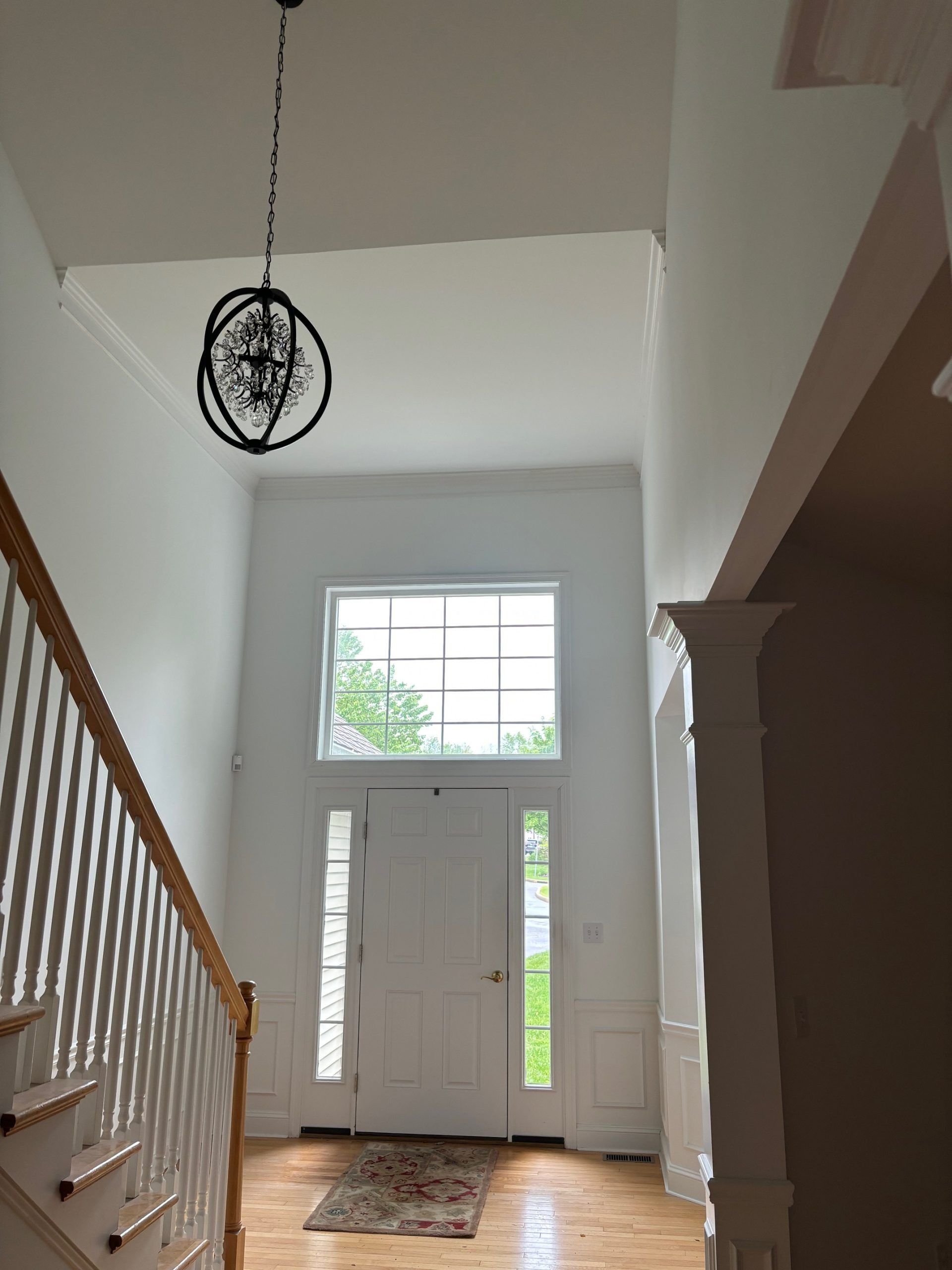 A bright, high-ceilinged foyer with a wooden staircase, a white front door with side panels, and a modern orb chandelier.