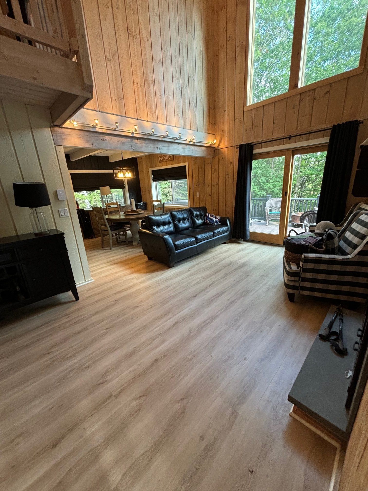 A spacious, rustic living room with wood paneled walls, high ceilings, a black leather sofa, and a large windowed wall.