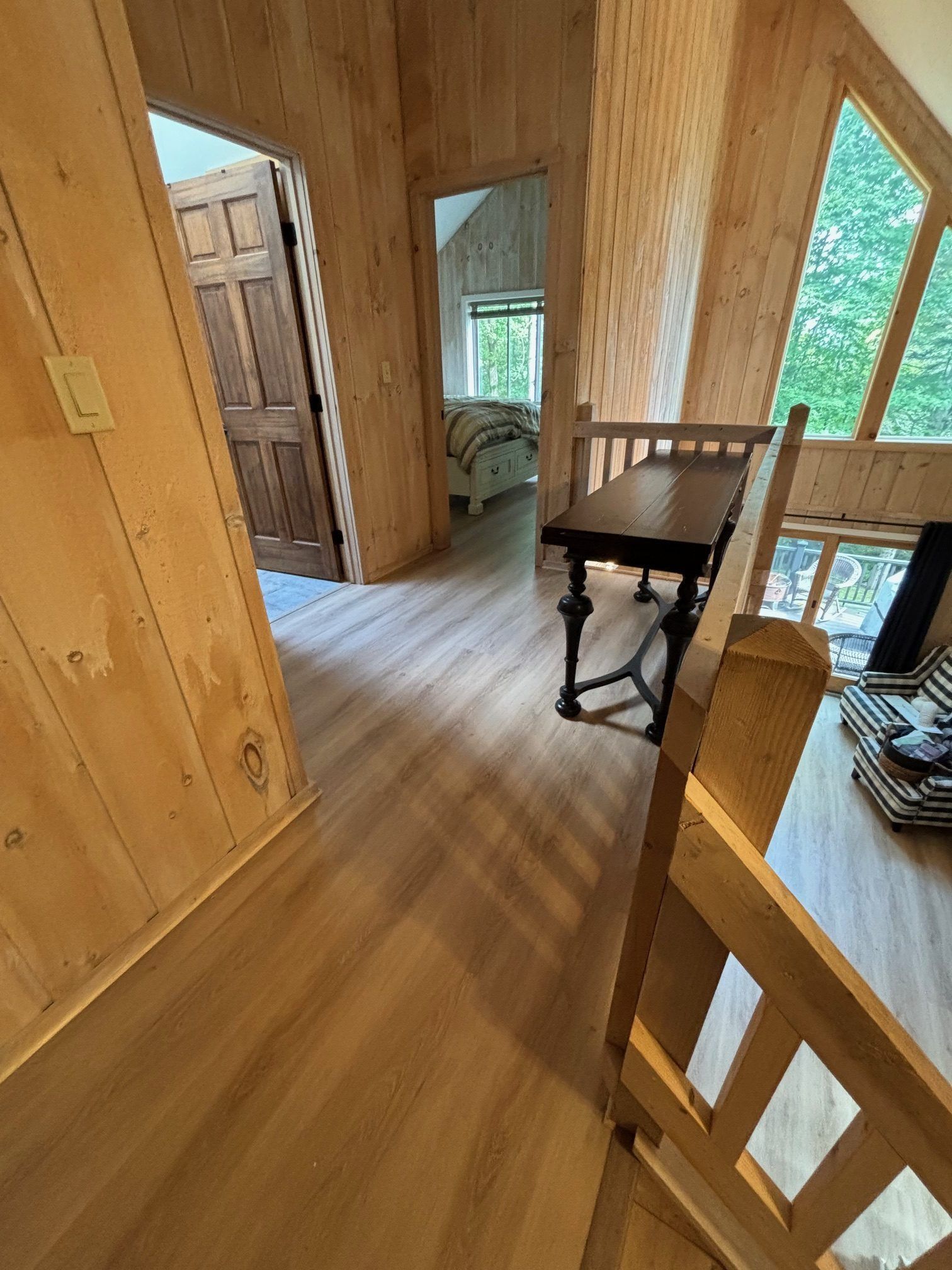 Wooden hallway with plank walls and flooring, leading to two bedroom doorways, with a wooden table and stair railing.