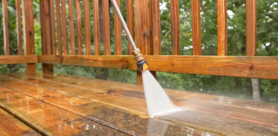 A pressure washer sprays water on a wooden deck, cleaning a strip of surface while removing dirt and discoloration.