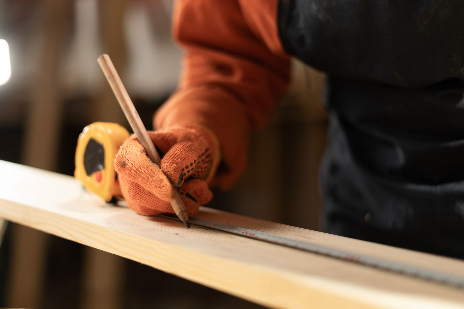 A person wearing orange work gloves uses a pencil and a tape measure to mark a line on a wooden plank in a workshop.