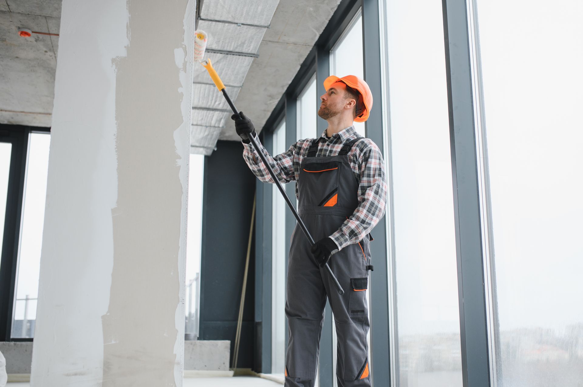 A construction worker in a hard hat and overalls uses a long-handled paint roller on a ceiling in a modern building.