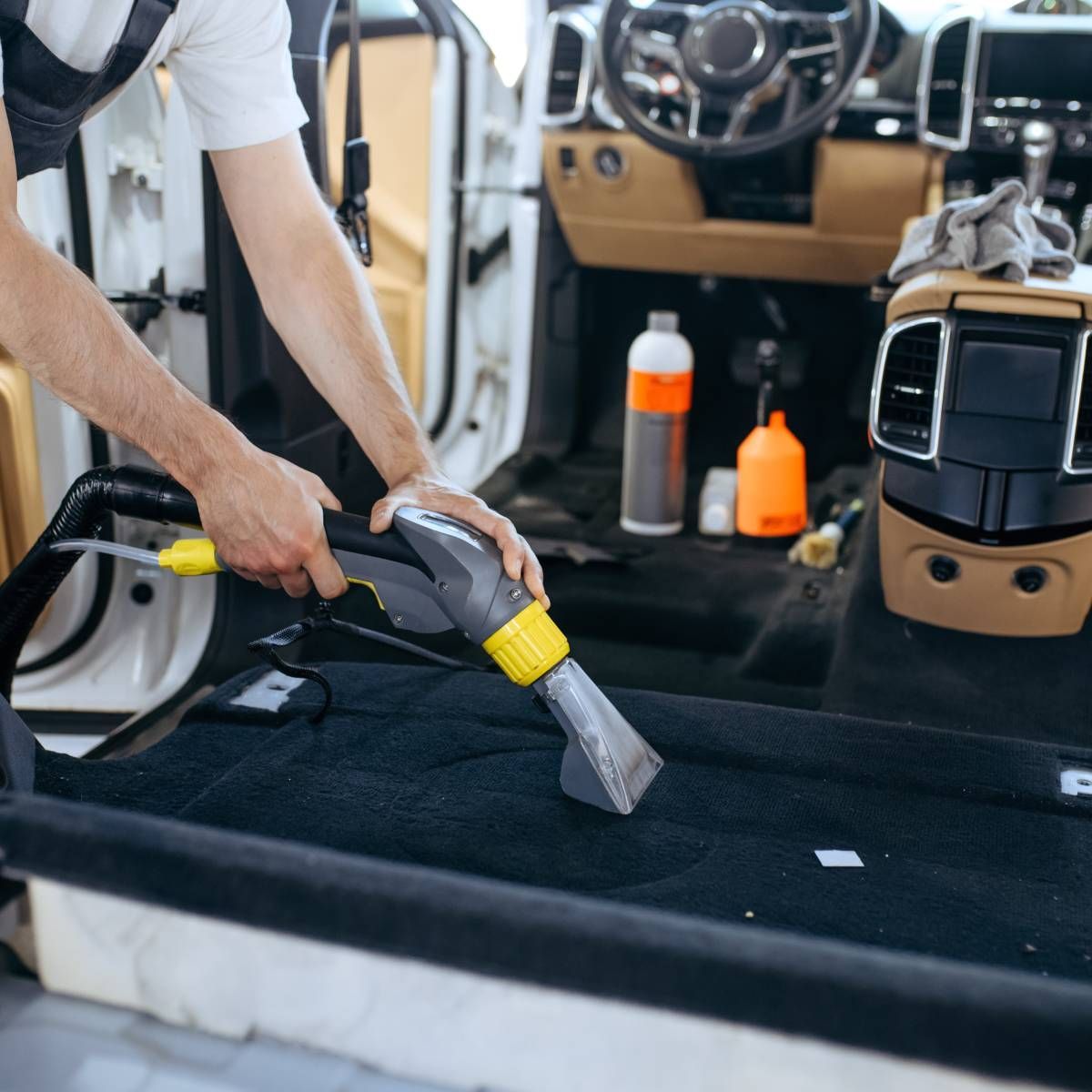 Person vacuuming a car's black carpet interior, using a handheld vacuum tool.