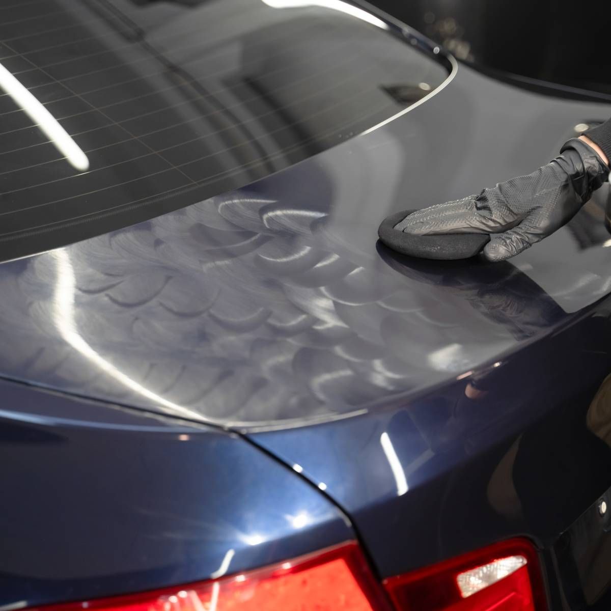 A person polishing a dark blue car with a gloved hand, showing swirl marks.