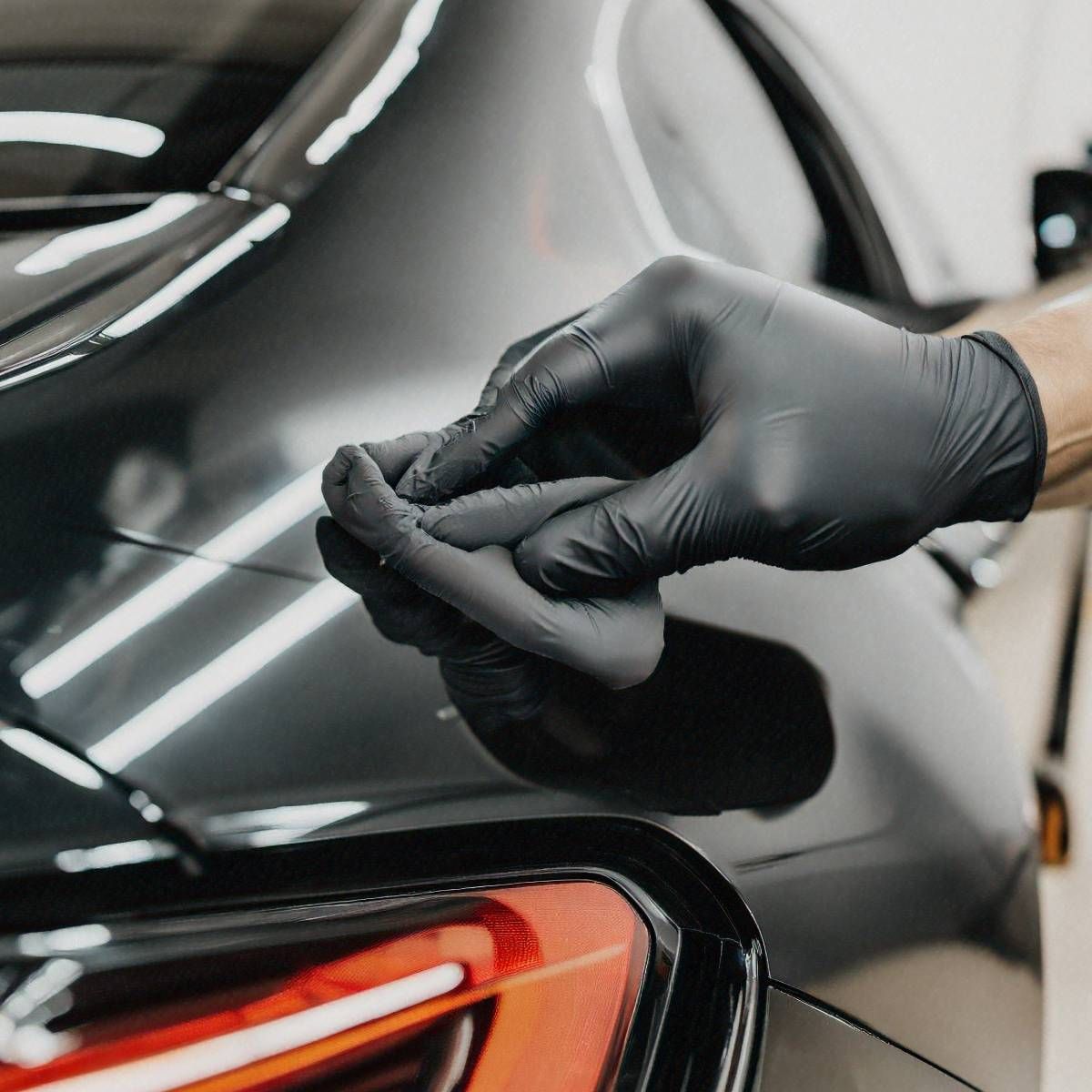 Gloved hand detailing a black car's rear panel. The hand applies product to the shiny surface near a red taillight.