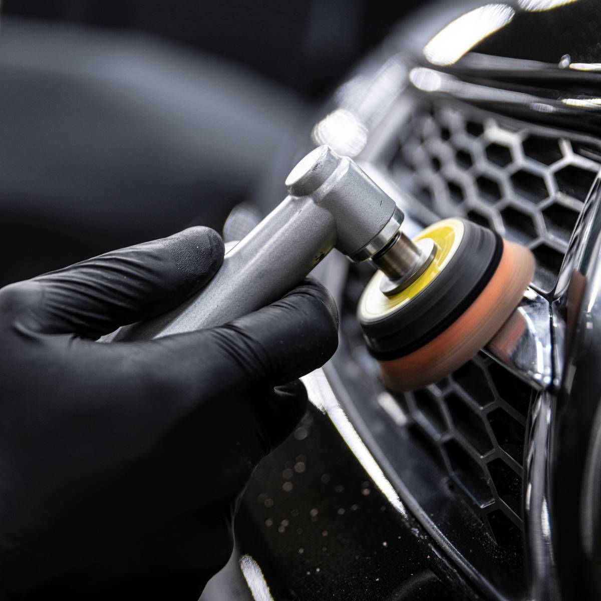 Black-gloved hand using a rotary polisher on a car's chrome grill, buffing and detailing.