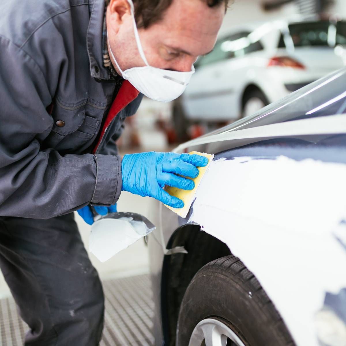 Mechanic in overalls and mask sanding a car fender.