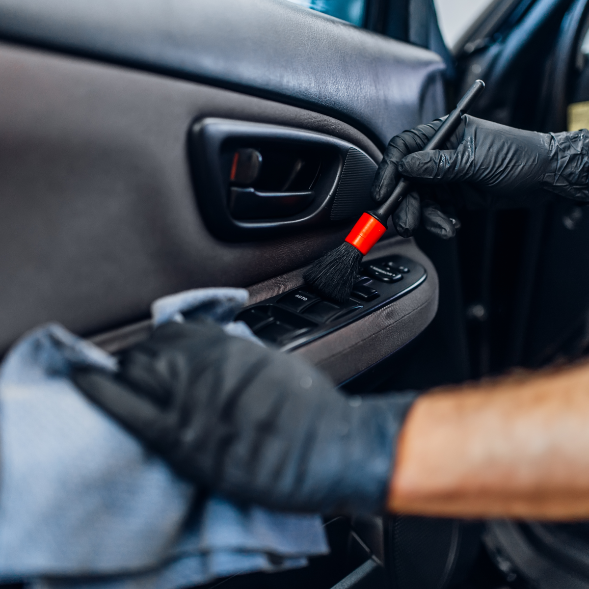 Person cleaning a car's interior door panel with a brush and cloth, wearing black gloves.