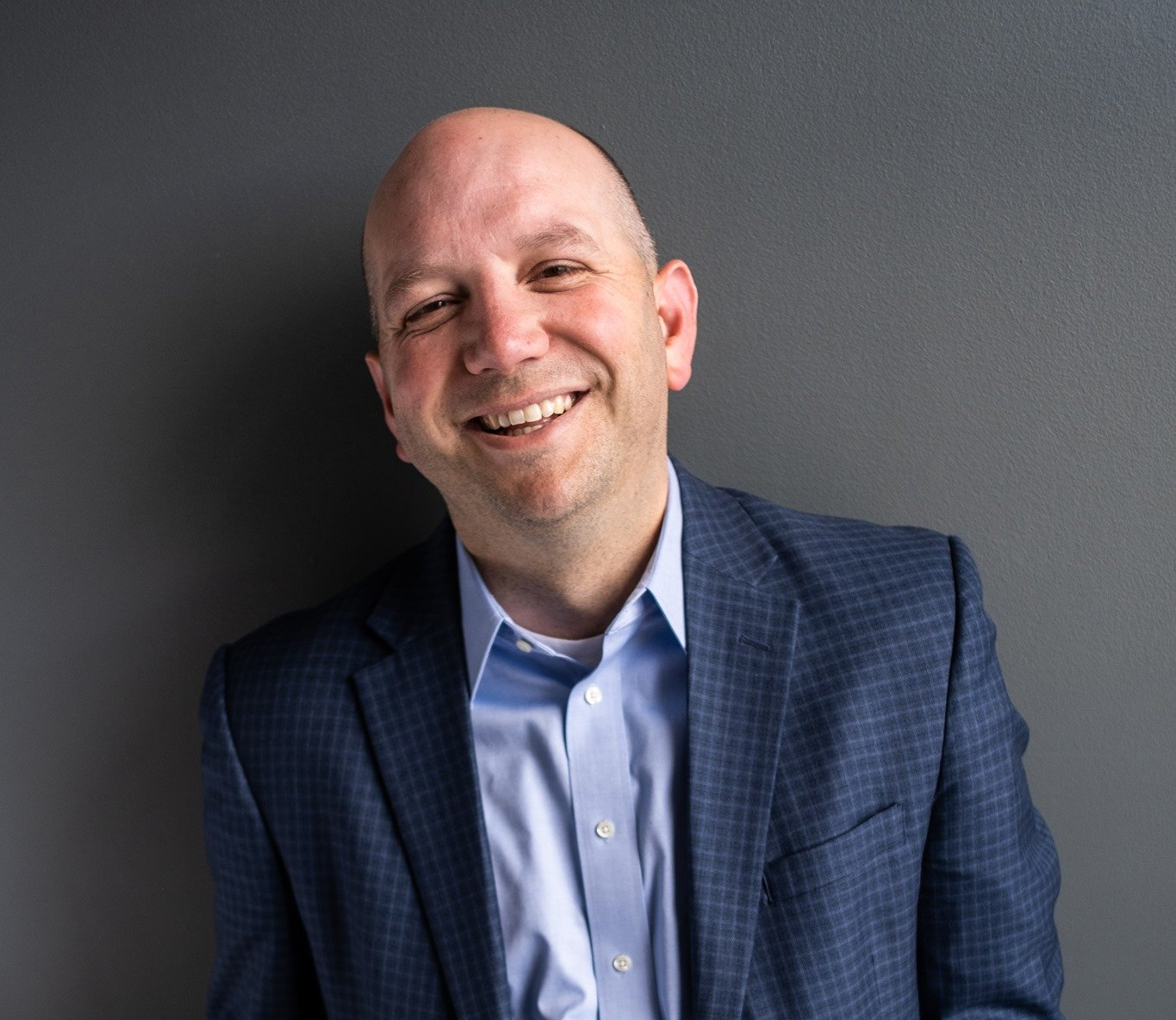 A man in a suit and blue shirt is smiling in front of a gray wall.
