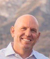 A bald man in a white shirt is smiling in front of a mountain.