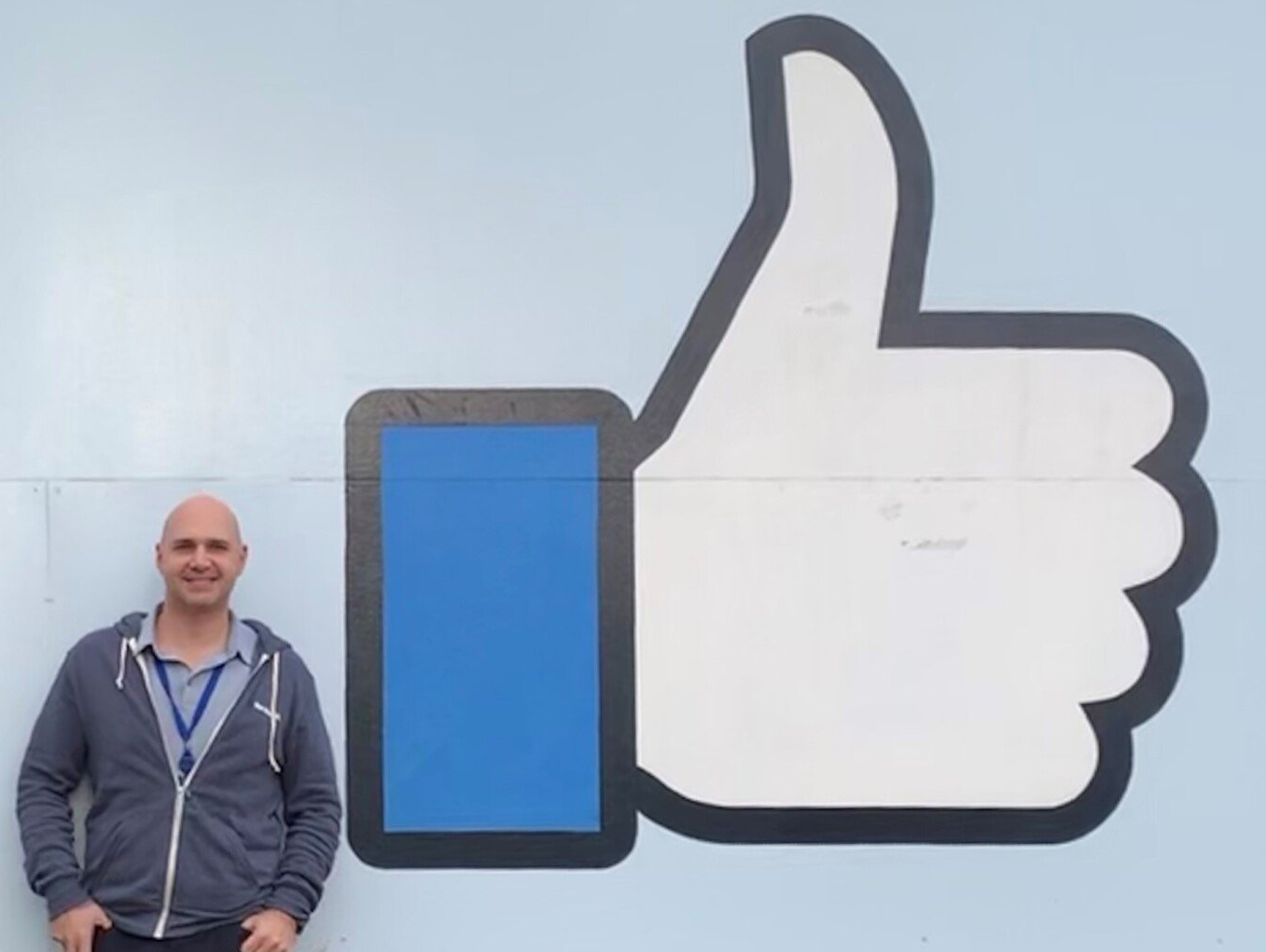 A man stands in front of a large facebook thumbs up sign