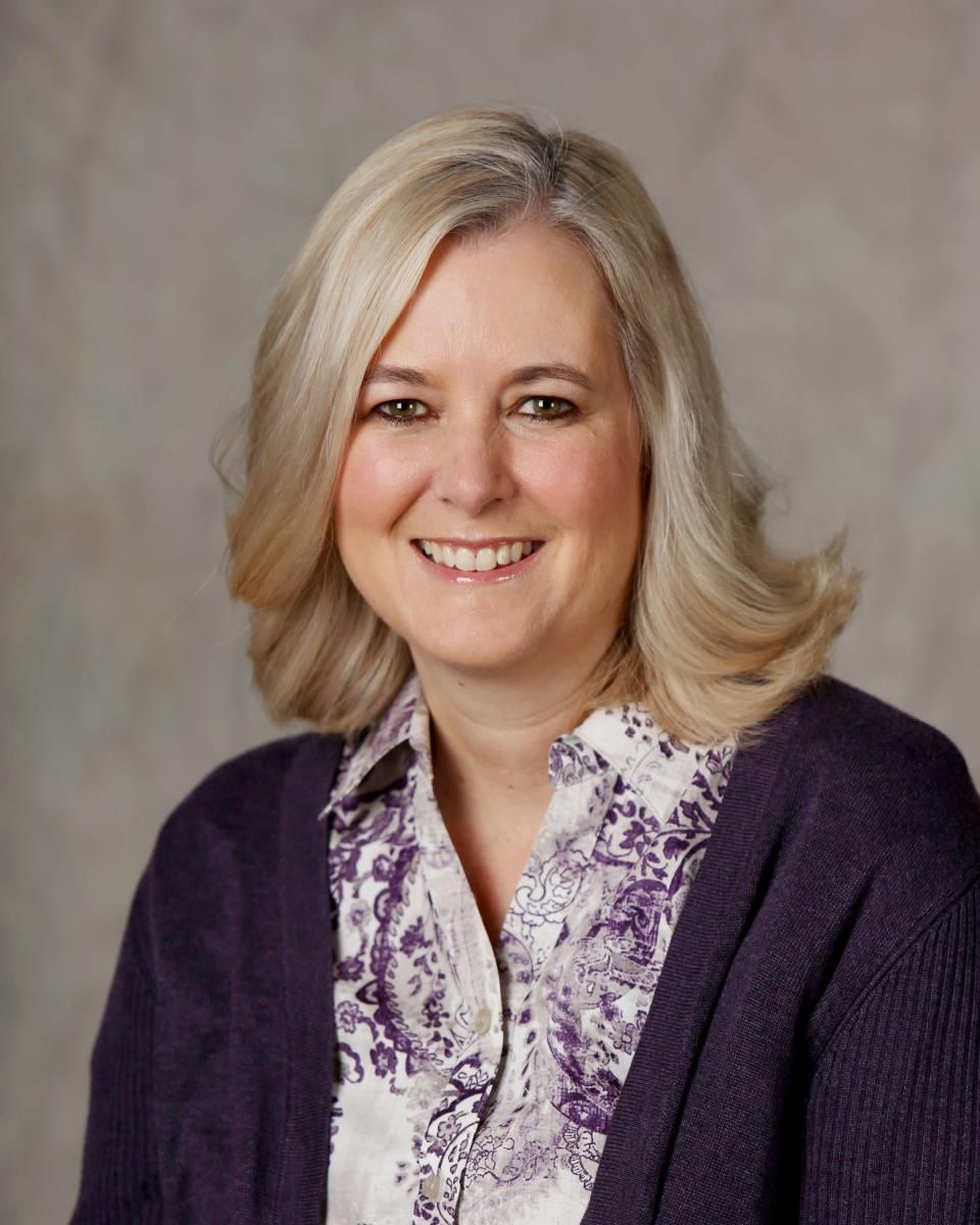 A woman wearing a purple sweater and a floral shirt is smiling for the camera.