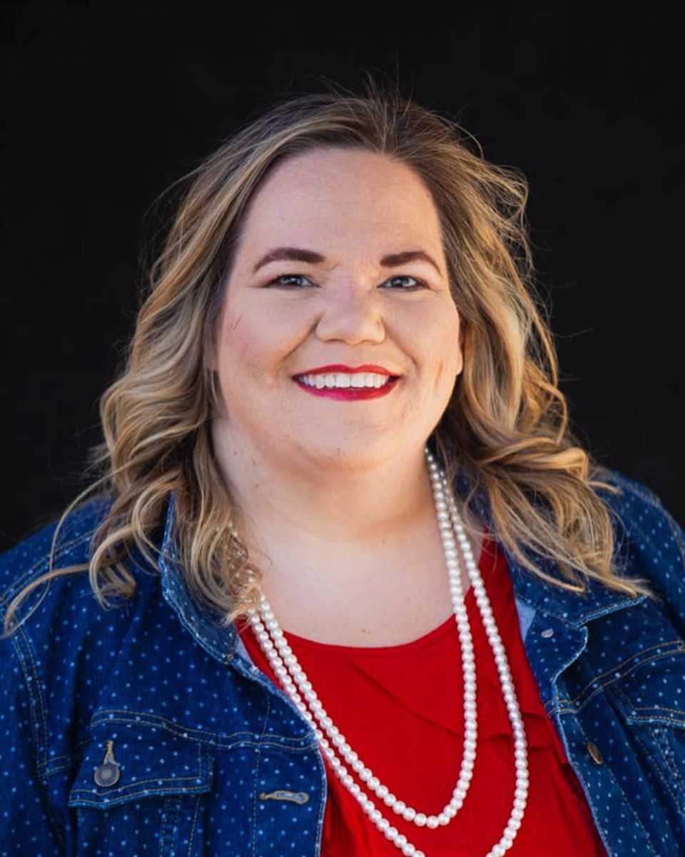 A woman wearing a red shirt and a pearl necklace is smiling for the camera.