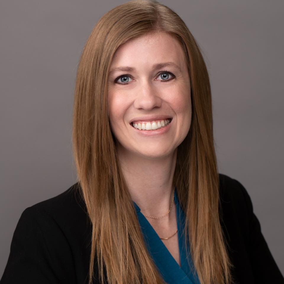 A woman with long brown hair is smiling for the camera while wearing a black jacket and a blue shirt.
