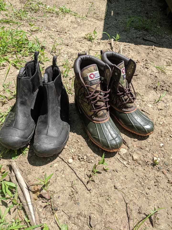 Two pairs of boots on dirt ground. One pair is black, ankle-high. The other pair is green and brown, mid-calf height