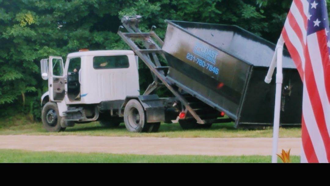 White truck with a dumpster lifting in front of trees and American flag.