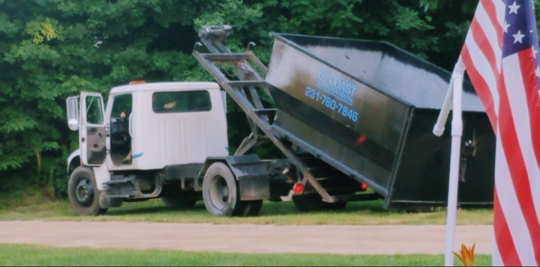 White roll-off truck with a black dumpster. The dumpster is raised. An American flag is visible.