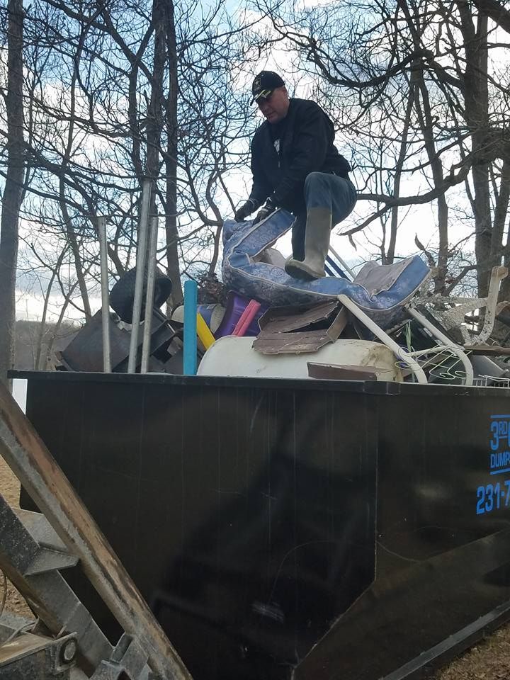 Man standing in dumpster, placing items inside. Outdoor setting, trees in background, blue sky.