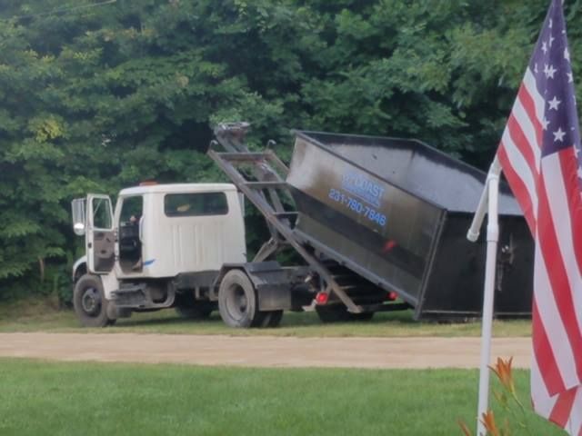 White truck lifting black dumpster on a grassy area, American flag in foreground, trees in background.