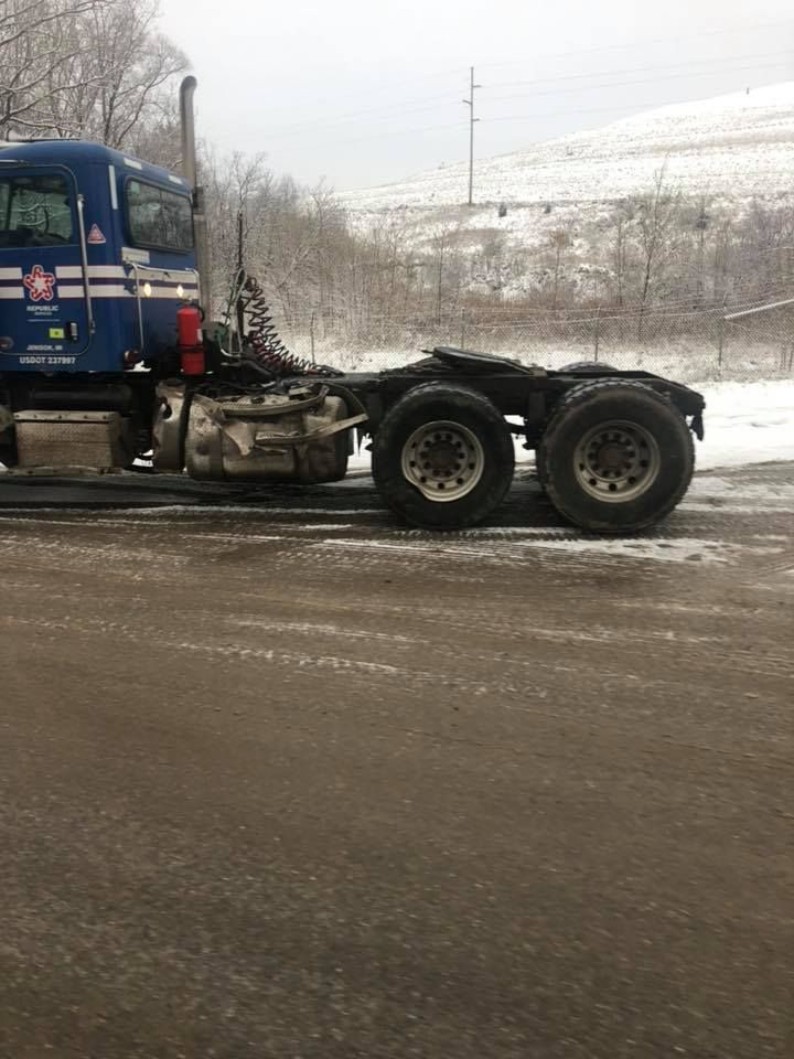 Blue semi-truck cab and chassis parked on snowy asphalt with a snow-covered hill in the background.