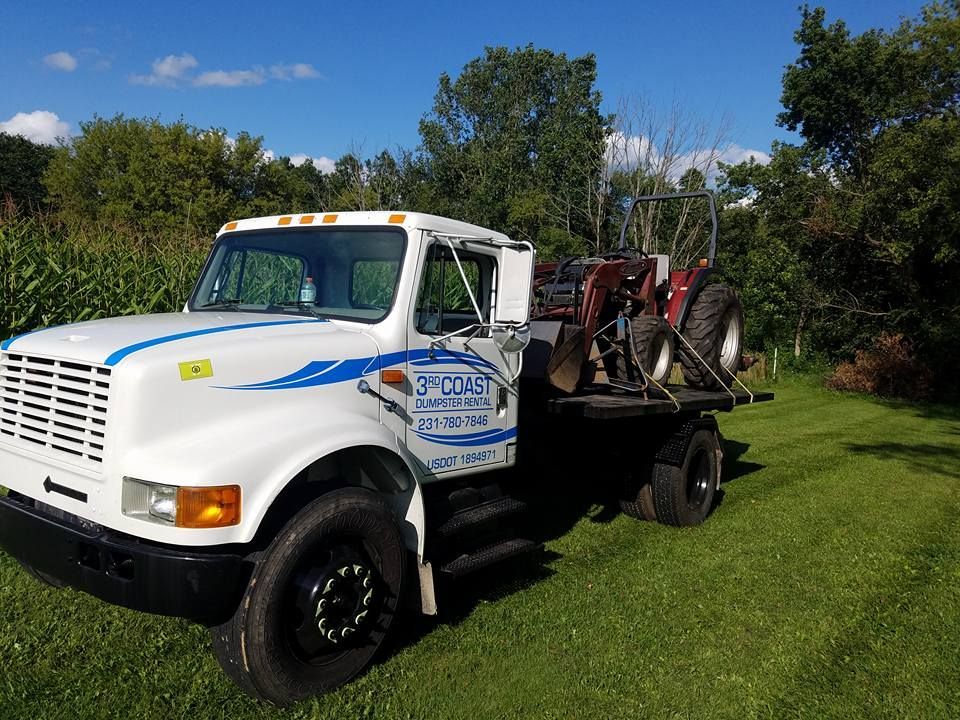 White truck with a flatbed carrying a red tractor on green grass with blue sky and trees.