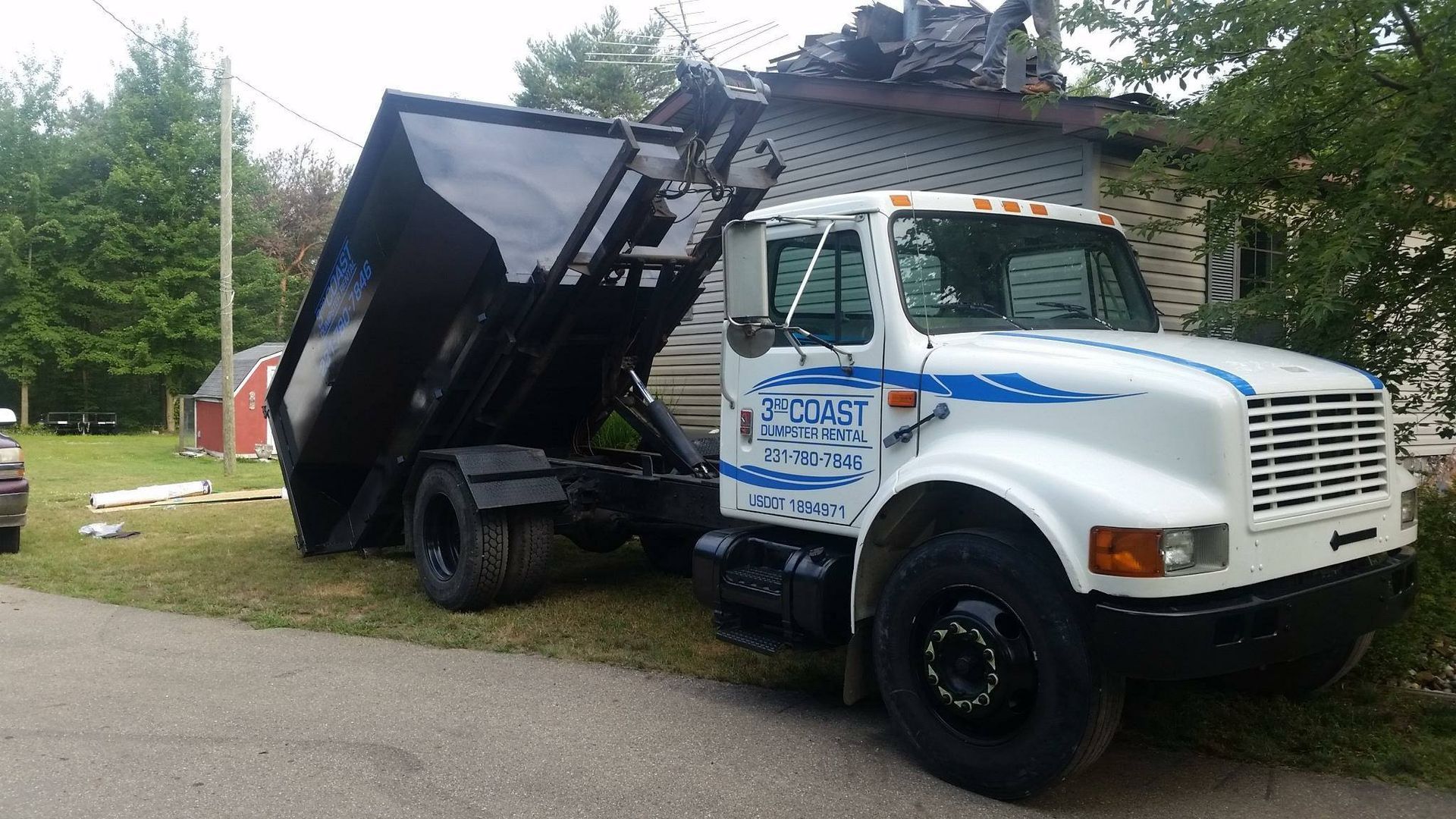 White dump truck with a raised black bed parked next to a house with debris on the roof.