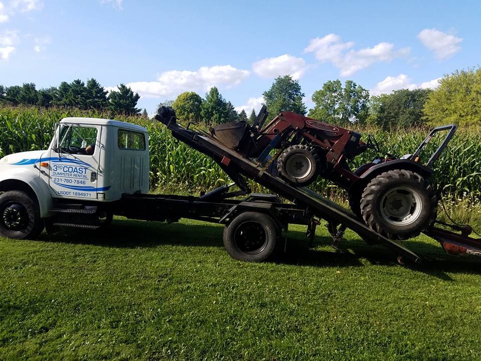 Tow truck transporting a red tractor on a sunny day in a grassy field next to a cornfield.