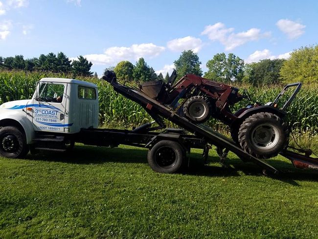 Tow truck transporting a red tractor on a sunny day in a grassy field next to a cornfield.