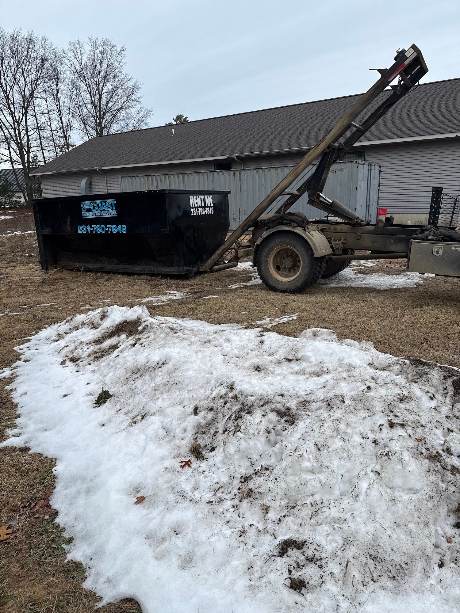 Black dumpster attached to a trailer, next to a pile of melting snow, in front of a building.