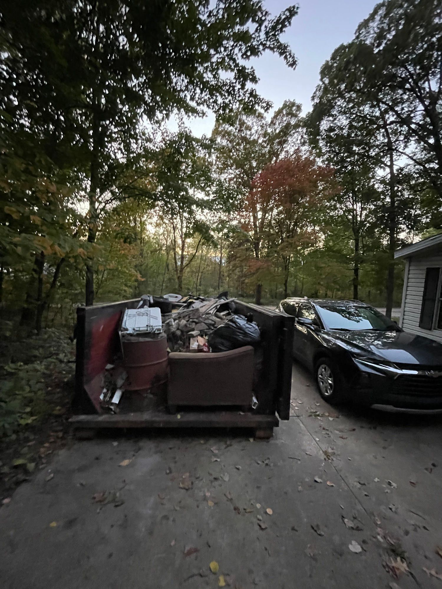 Dumpster overflowing with trash, next to a black car and a house in a wooded area.