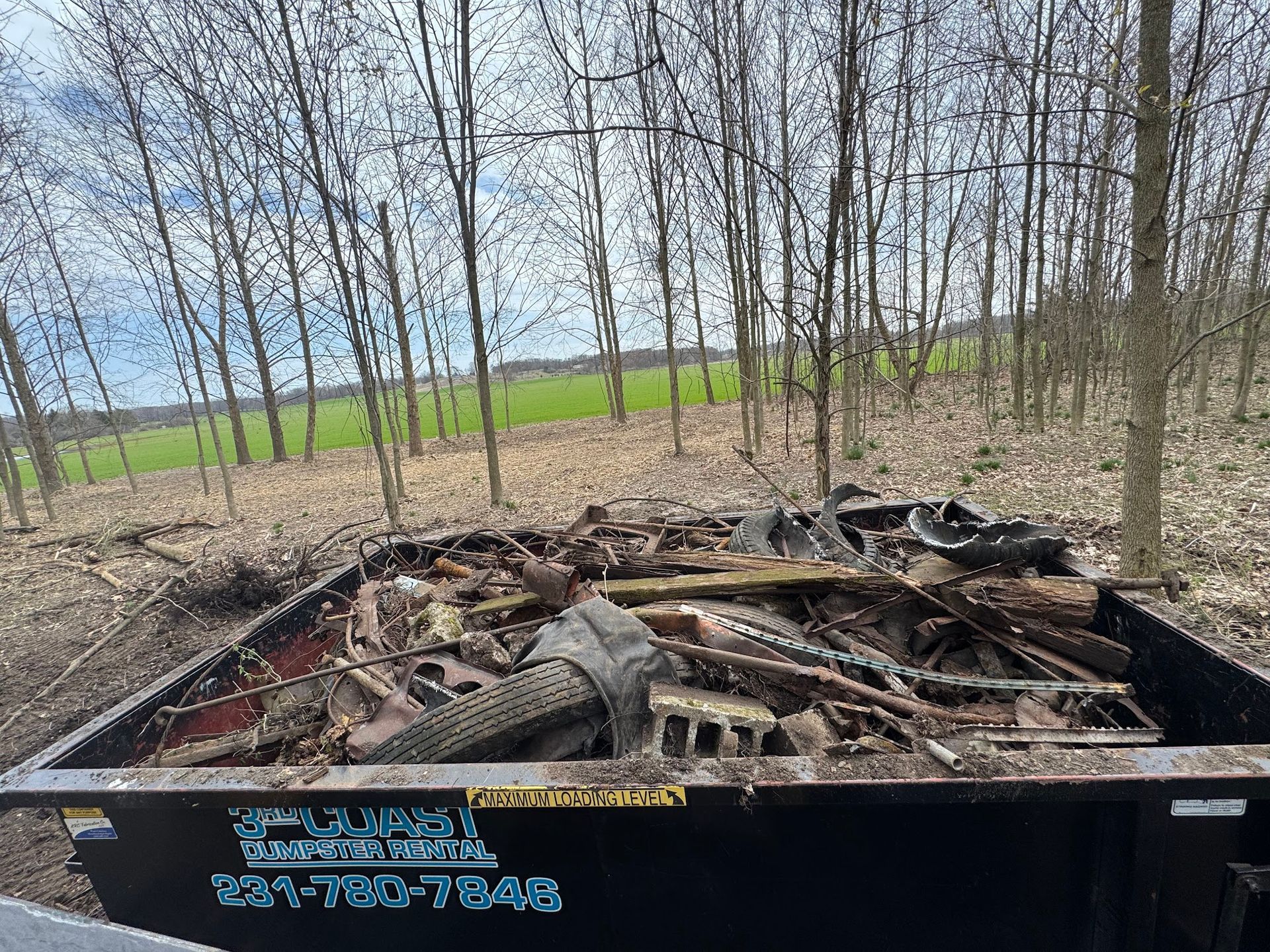 A full black dumpster filled with construction debris sits in a wooded area with a field in the background.
