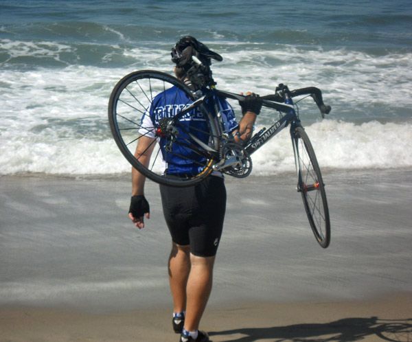 A person carrying a bicycle on the beach with a shirt that says fortuna