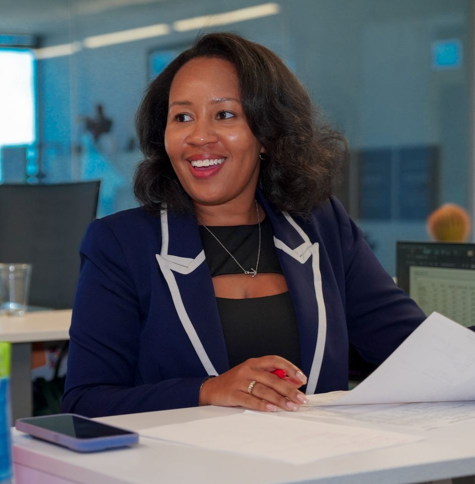 Woman in a blazer on a phone, smiling while using a laptop at a wooden desk in an office.