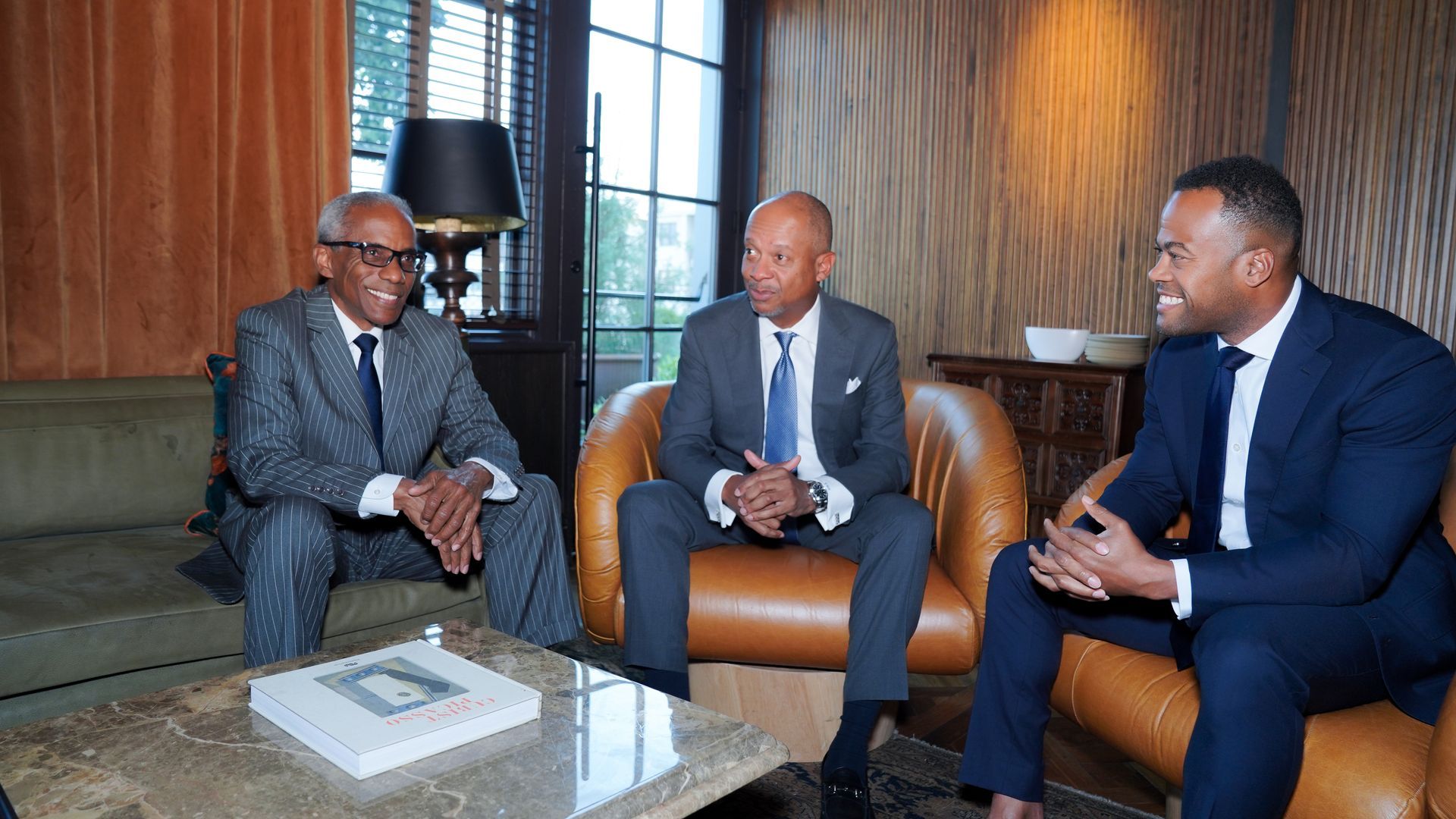 Three people in business suits sit in armchairs in a well-lit lounge area, engaged in conversation.