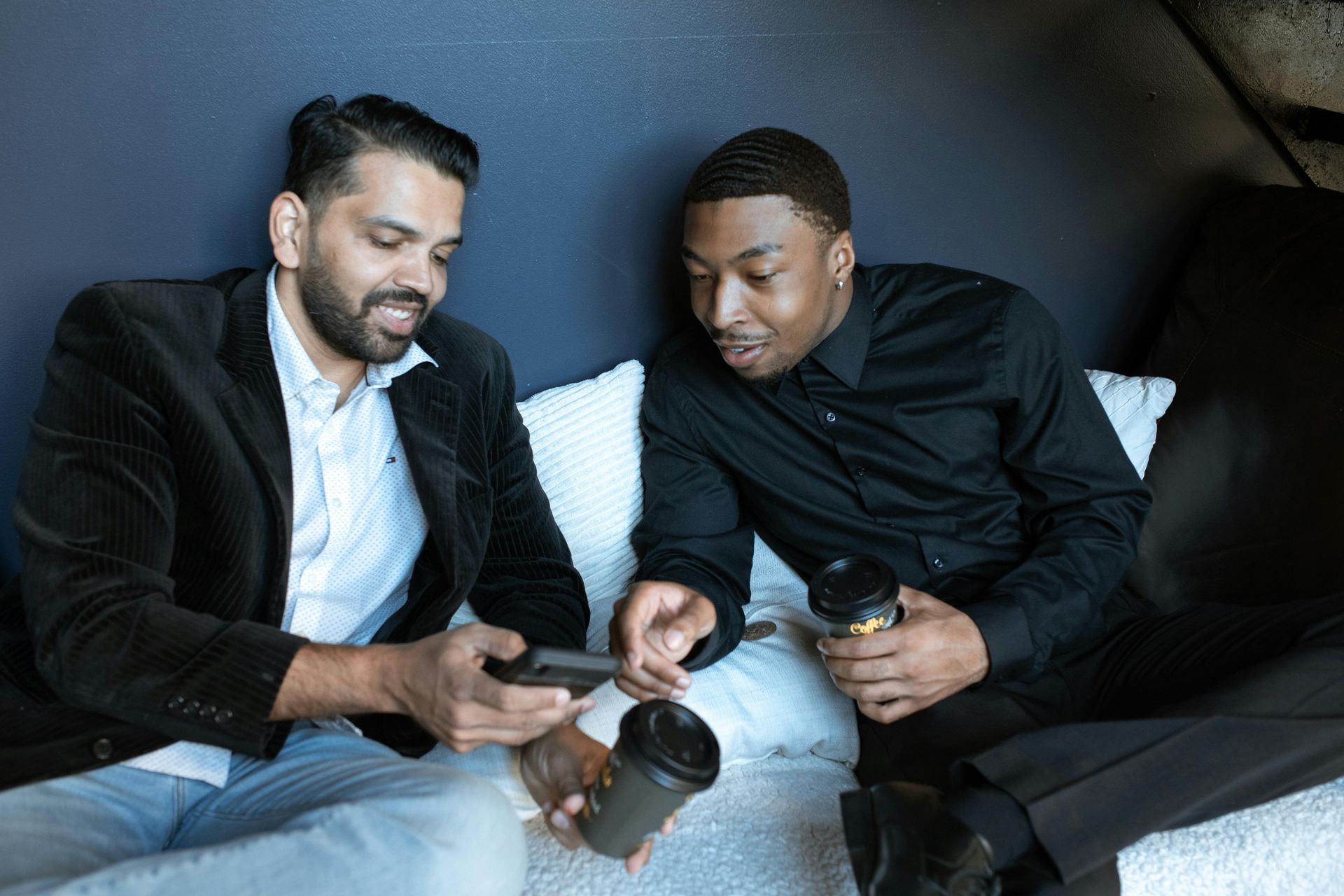 Two men are sitting on a couch looking at a cell phone.