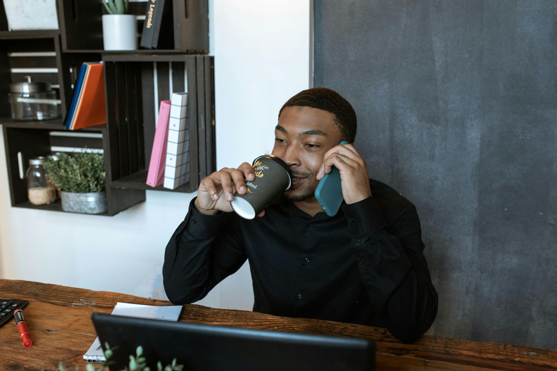 A man is sitting at a desk drinking coffee and talking on a cell phone.